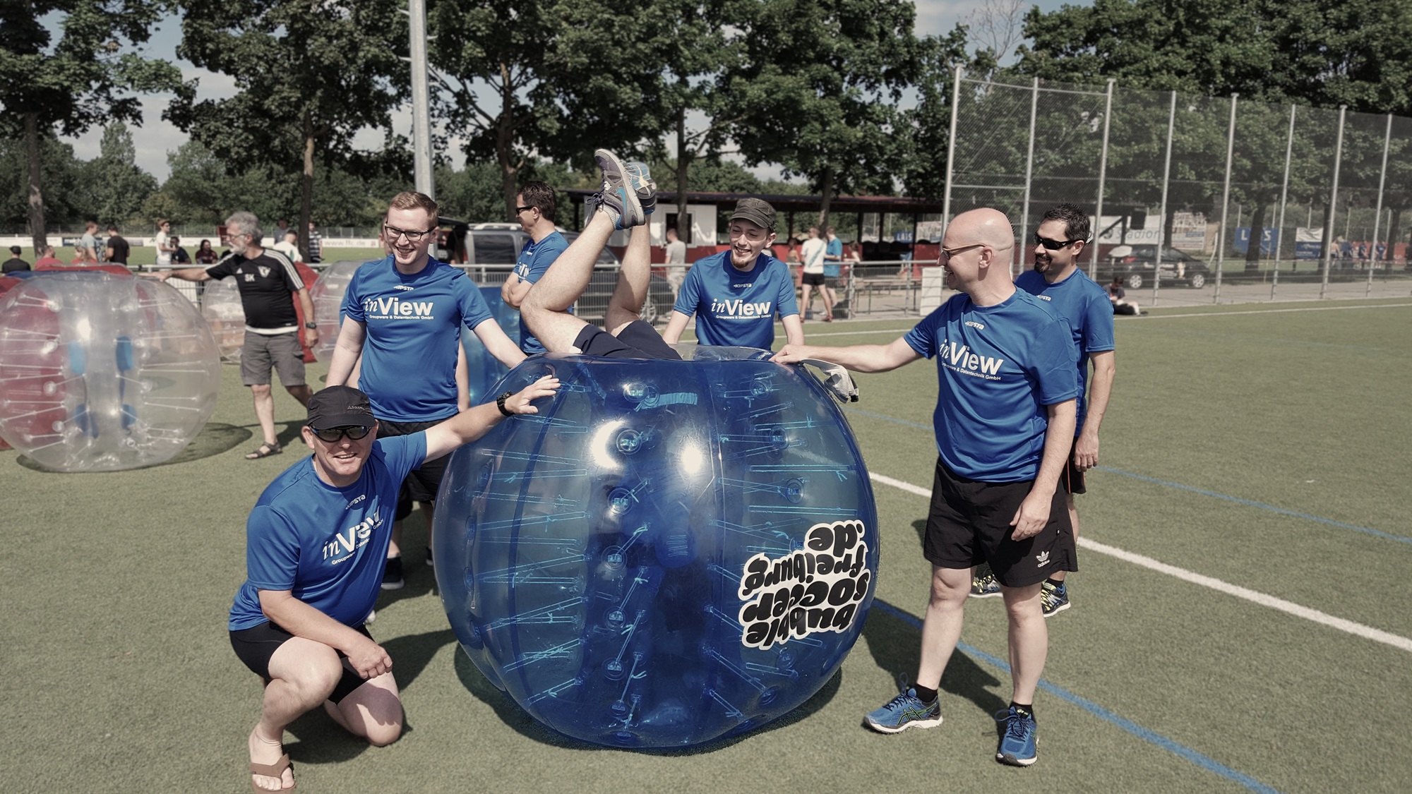 inView Groupware und Datentechnik GmbH Team in blauen Shirts beim Bubble Soccer. Ein Mitarbeiter steckt in einem großen Ball umkreist von den anderen Teammitglieder – Symbol für Zusammenhalt, Teamgeist und eine offene Unternehmenskultur im IT-Bereich