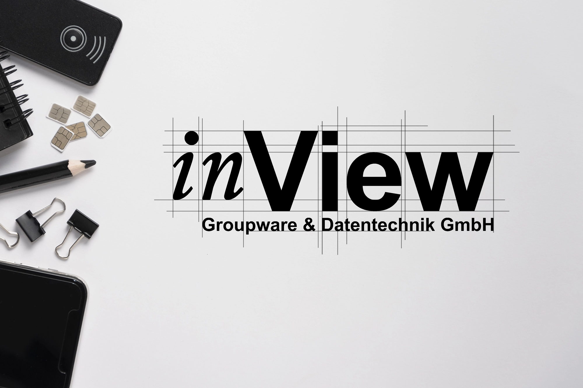 Weißer Arbeitsbereich mit Handy, Bleistift, Binderklammern und SIM-Karten auf der linken Seite; auffälliges schwarzes „inView“-Logo mit Gitterlinien und der kleinen Unterschrift „Groupware & Datentechnik GmbH“ mittig rechts, passend zur ganzheitlichen IT-Betreuung und modernen Kollaborationslösungen eines IT-Dienstleisters aus Freiburg